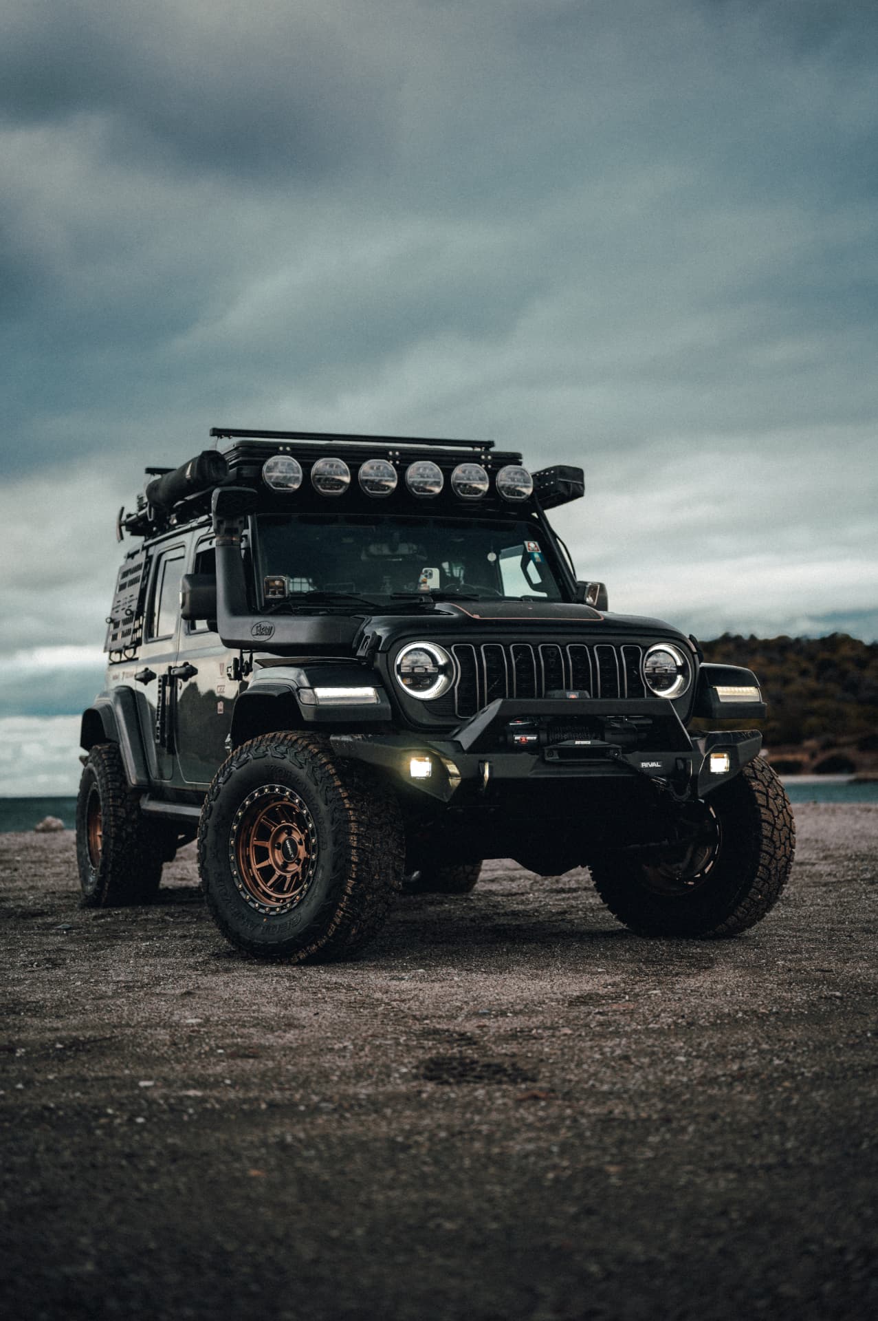 Black Jeep Wrangler with roof-mounted light bar and bronze wheels on a gravel shore under an overcast sky